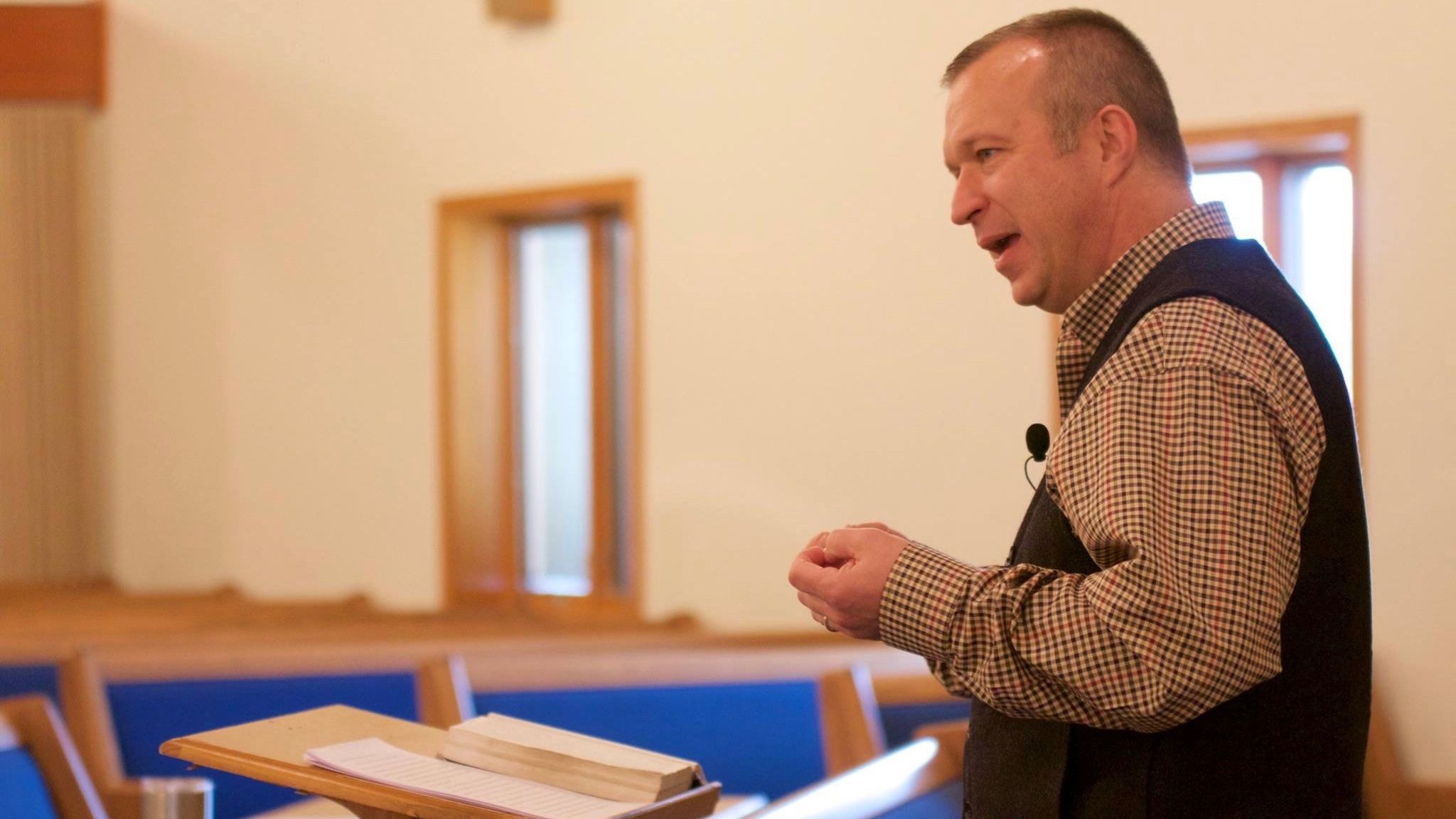 A speaker in a plaid shirt and vest addresses a congregation from a wooden pulpit