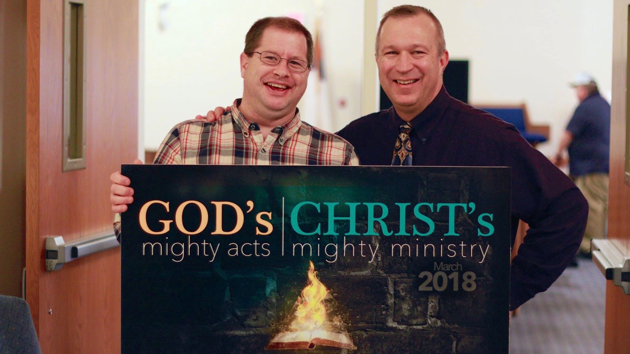 Two men hold a banner reading “God’s mighty acts, Christ’s mighty ministry 2018”